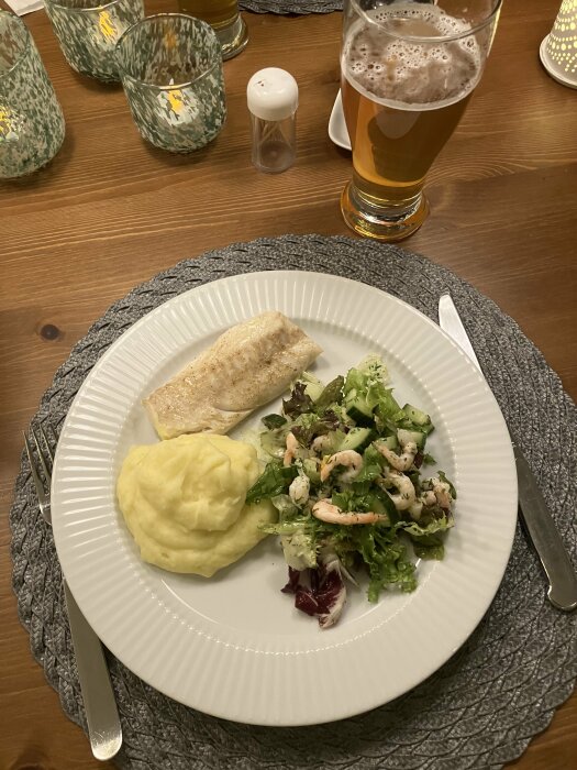 En måltid med fisk, potatismos, räksallad, ölglas, saltkar och ljusa tallrikar på ett träbord.