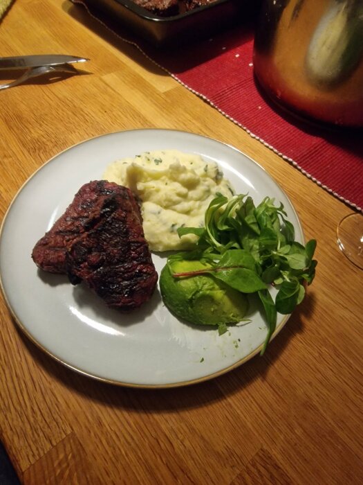 Grillad steak, mosad potatis med persilja, spenatsallad och skivad avokado på en tallrik.