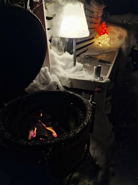 Eldstad utomhus i snö med brinnande ved, omgiven av snöklädda ytor och julbelysning.