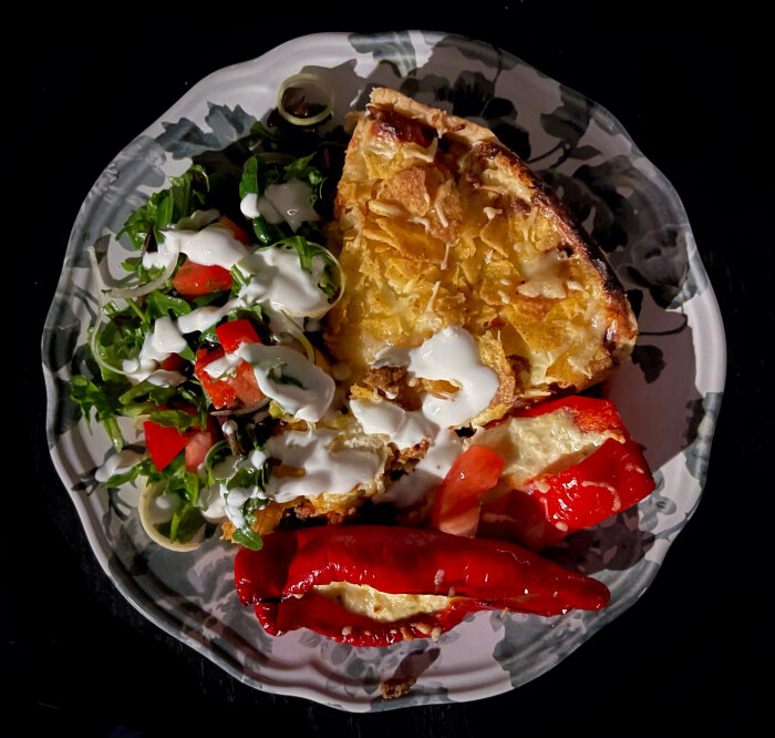Ett fat med paj, gräddfil, sallad, tomater, rucola och röd paprika, mörk bakgrund.