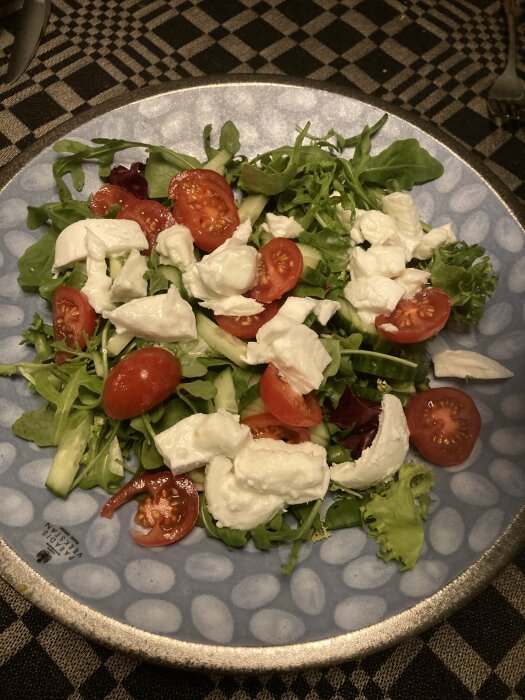 En sallad med blandade gröna blad, tomat och mozzarella på ett blå-vitt mönstrat fat.