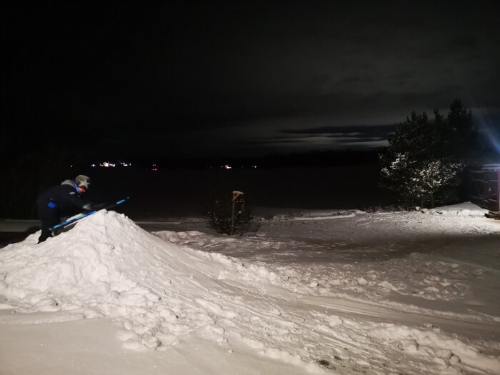 Person åker nerför snöbacke på natt, ljus från hus belyser landskapet i bakgrunden.