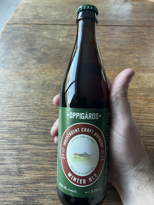 Hand håller en Oppigårds Winter Ale flaska ovanför ett träbord.