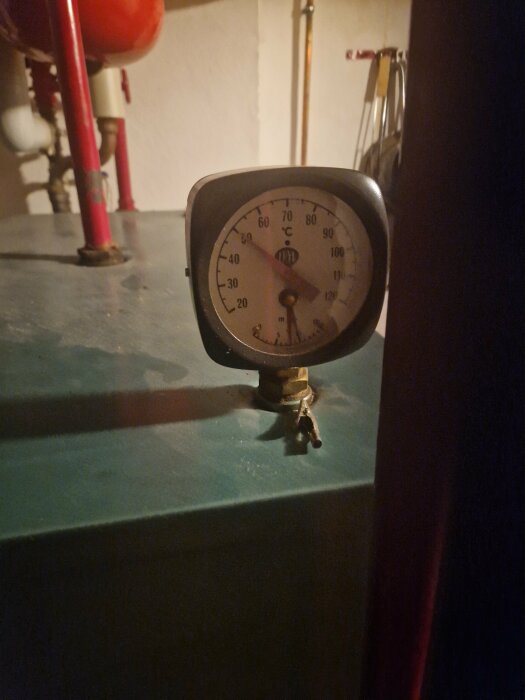 Analog termometer på en grön yta, industriell miljö, rörledningar i bakgrunden, svagt upplyst, temperatur cirka 60 grader Celsius.