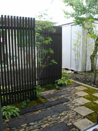 Trädgårdspassage med staket, mossbelagd stenstig, växter, och dekorativt grus. Utomhus, lugnt, grönt.