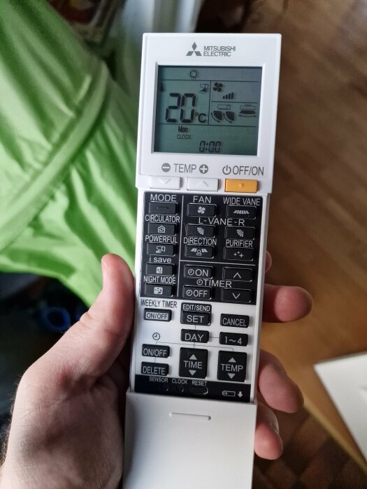 En hand håller en Mitsubishi Electric fjärrkontroll för AC med display som visar 20 grader Celsius och måndag 00:00.