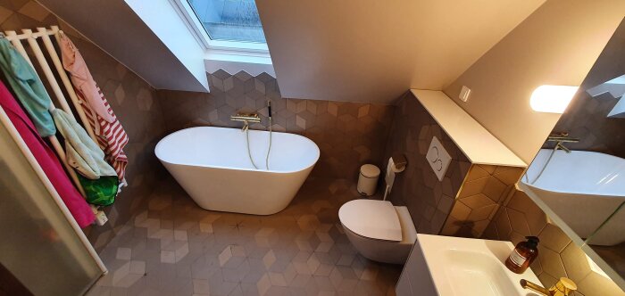 Modernt badrum med fritstående badkar, handdukstork, toalett, handfat, guldarmatur och takfönster.