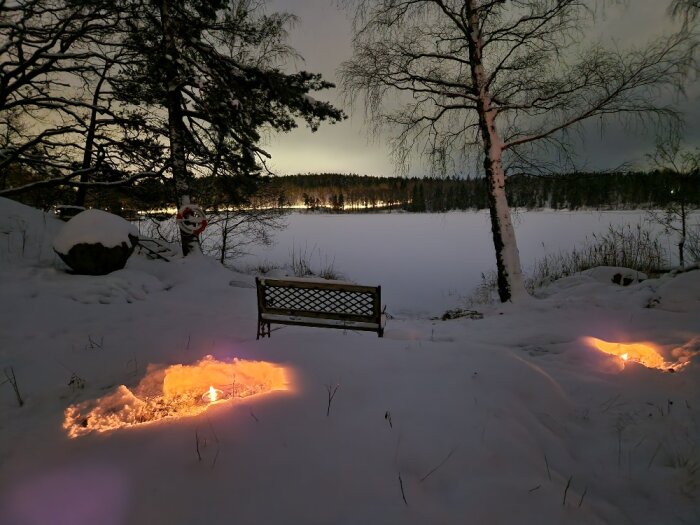 Vinterlandskap nattetid, snö, bänk, träd, belyst hål i marken, fruset vatten, molnig himmel.