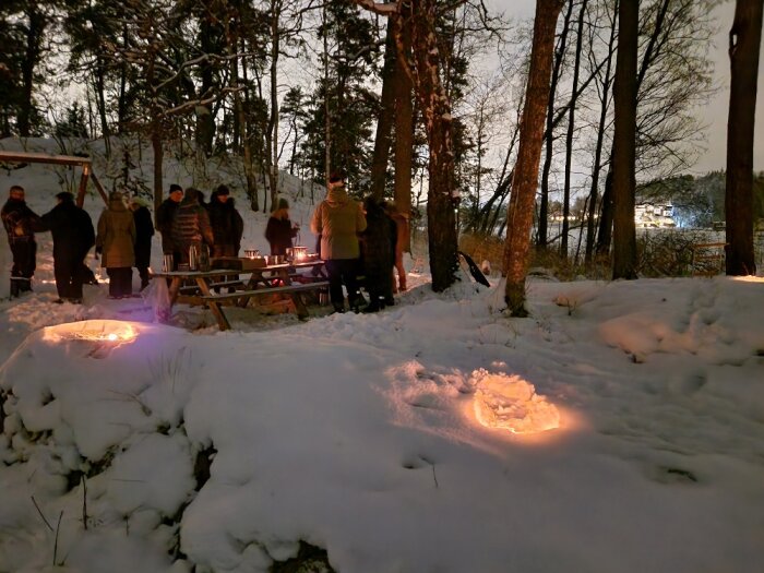 Vinter, snö, människor vid eldar, skymning, utomhus, samvaro, mörkt trädlandskap, varma ljuskällor, mysig stämning.