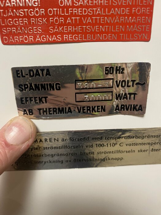 Etikett med teknisk information, finger håller, text på svenska, varningsetikett, elektriska specifikationer, sliten yta.