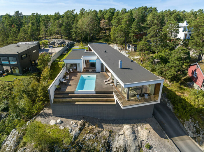 Modernt hus med terrass, pool och stora fönster, omgivet av skog och klippor, luftig vy.