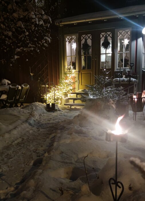 Kväll, snötäckt trädgård, upplysta fönster, juldekorationer, ljusstake utomhus, mysig vinterstämning, gångväg.