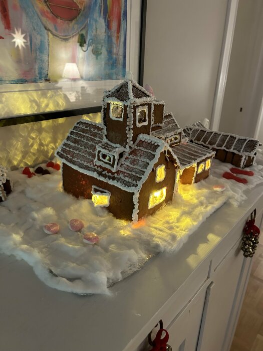 Ett pepparkakshus med belysning och strösockersnö, beläget på ett vitt bord med juldekorationer.