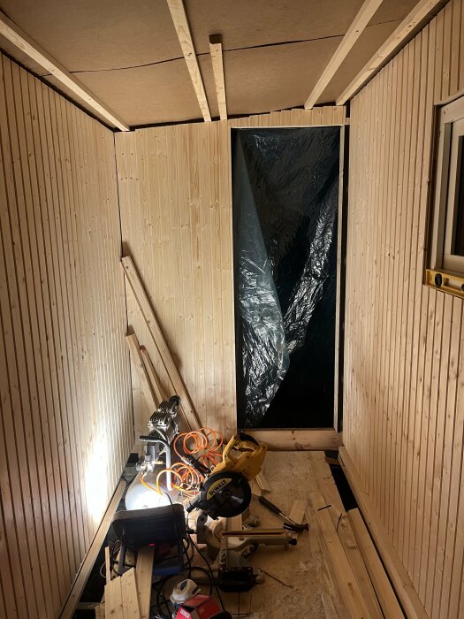 Rum under konstruktion med träpanel, verktyg och oisolerat isoleringsmaterial synligt. Byggkaos och träreglar.