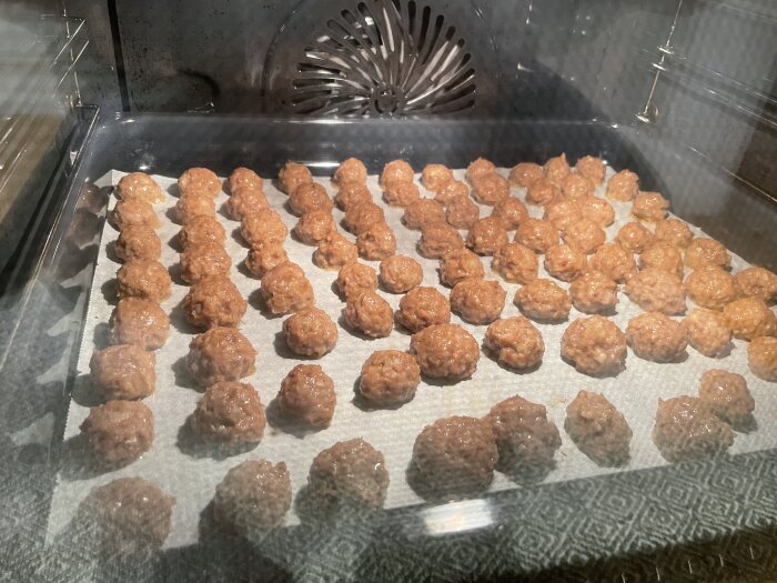 Råa köttbullar på bakplåtspapper i ugn, ordnade i rader, redo att tillagas.