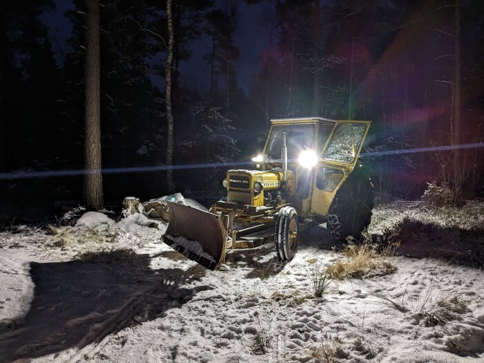 En gul grävmaskin i snötäckt skog, natt, strålkastare på, avger ett varmt ljus.