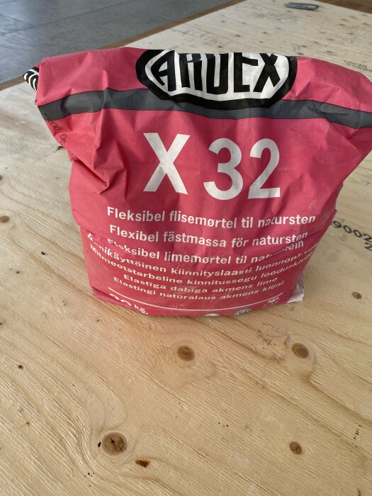 Rosa säck med text "Alfix X 32" på träyta. Byggmaterial, flexibel fästmassa för natursten, flisemørtel.
