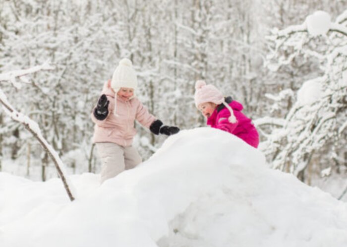 Två barn leker i snön, bygger snövall, vinterkläder, snöklädda träd i bakgrunden.