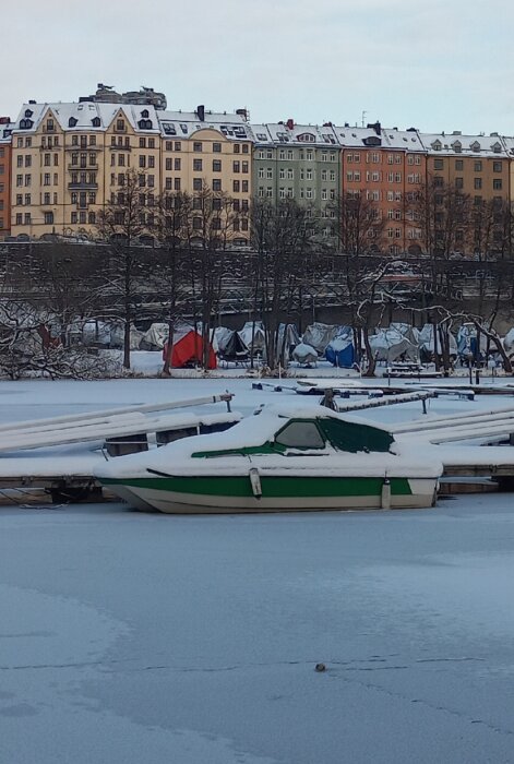 Båtar i isbelagd hamn, snö, färggranna byggnader i bakgrunden, vintertid, stilla vatten.