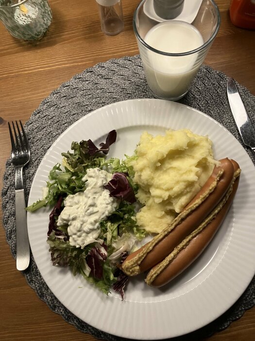 Middagsmåltid med korv, potatismos, sallad, dressing, glas mjölk och kryddor på träbord.