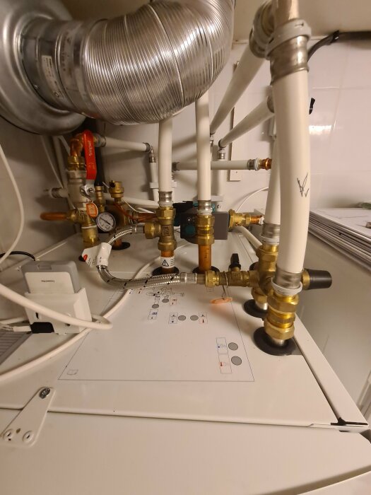 VVS-installation med rör, mätare och ventil ovanpå en vit apparat, möjligen en värmepanna eller liknande.