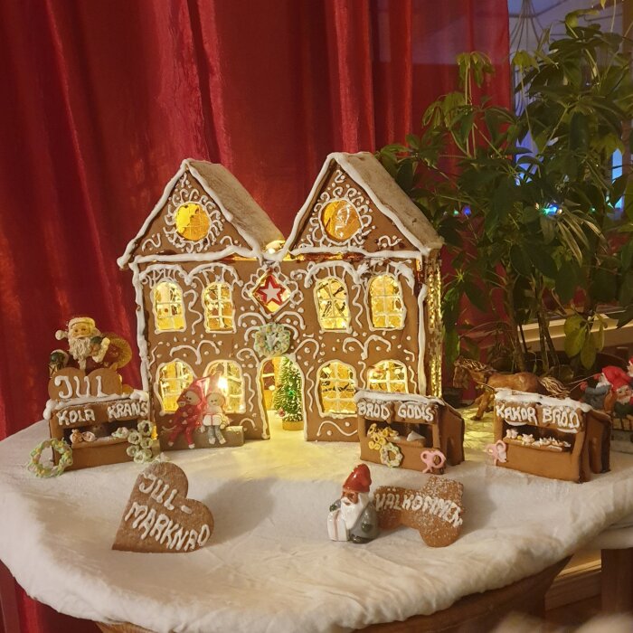 Pepparkakshus med dekorationer, figurer, röd bakgrund, julstämning, ljus från fönster, texter på svenska, "Julmarknad", miniatyrmarknadsstånd.