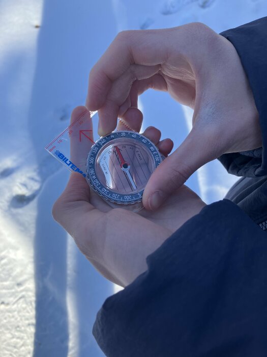 Händer håller kompass mot snöbakgrund, orientering, navigation, dagsljus, vinteraktivitet, utomhus.