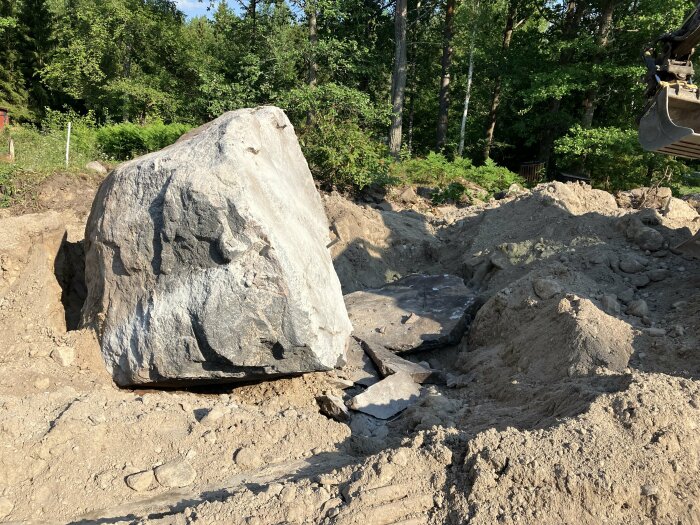 Stor sten på en grävplats, grävmaskinsskopa synlig, skog i bakgrunden.