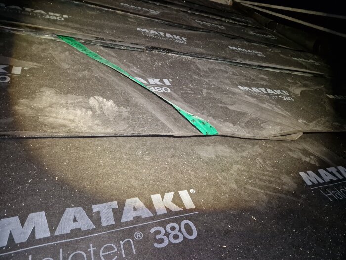 Mörk bild på takpapp med MATAKI-logotyp och grön rem överlagrade lager. Smuts och byggmaterial synliga.