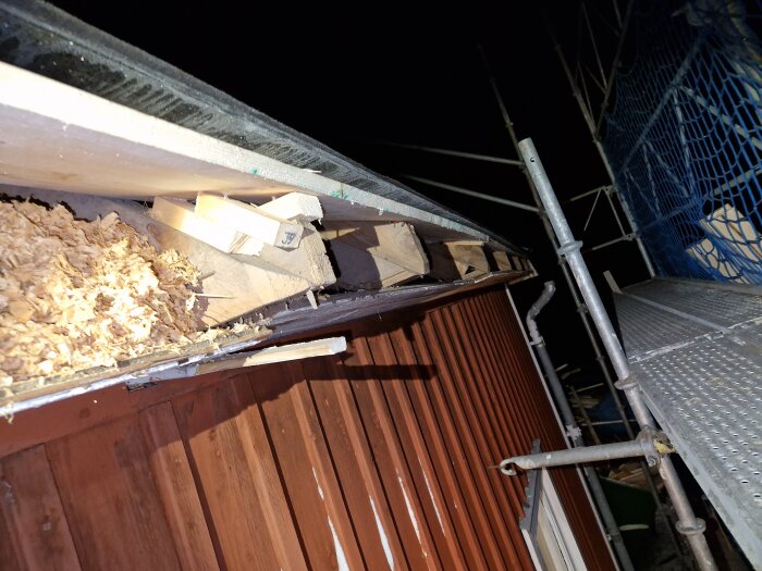 Byggnadsställning vid hus med öppen takkonstruktion och synliga isoleringsmaterial på natten.