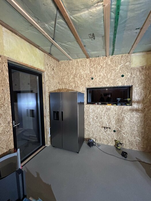 Utomslutande byggnadsarbete, OSB-paneler på väggar, isolering i tak, kylskåp, inbyggd TV, byggplats, inomhusmiljö.