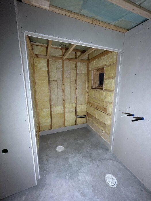 Ett oavslutat rum med isolering, gipsskivor, betonggolv och en liten fönsteröppning. Byggprocess eller renovering pågår.