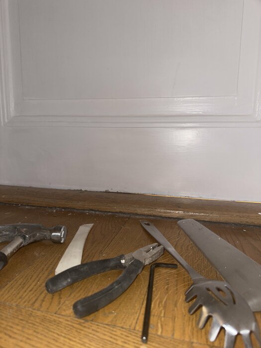 Verktyg på golv: hammare, mejsel, tång, såg och spik, vid en dörrlist. Hantverksarbete pågår, inomhus.