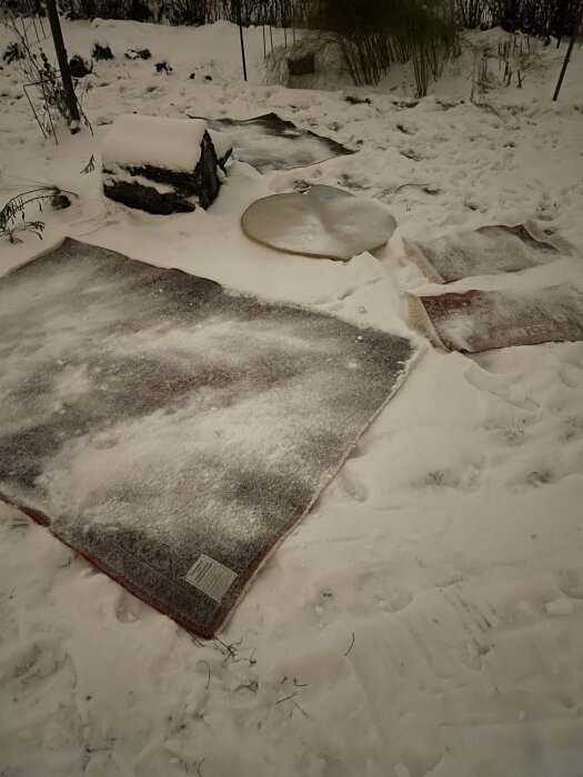 Mattor och en bordsskiva täckta av snö i en trädgård på kvällen.