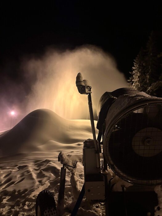 Snökanon skapar snötäcke nattetid på skidbacke med belysning och trädbakgrund.
