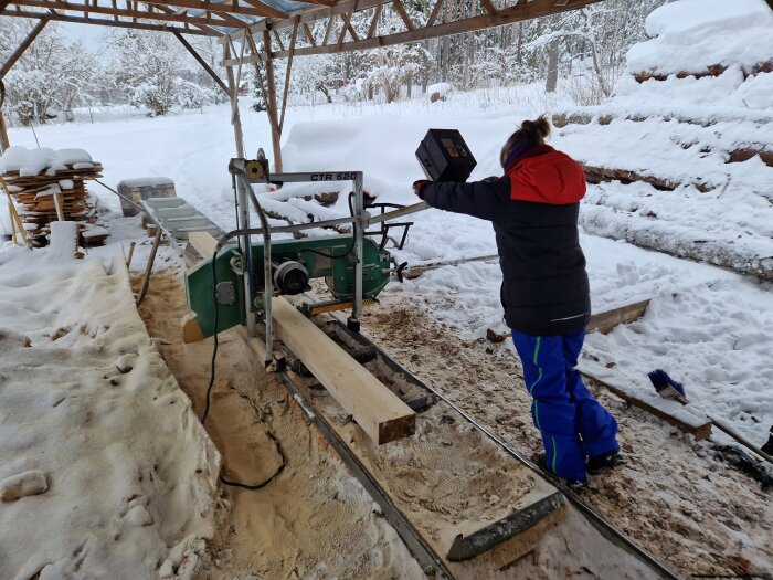 Person bedriver sågning med bandsågverk utomhus i snötäckt miljö.