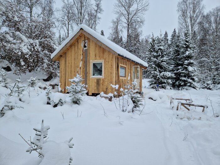 Ett litet trähus omgivet av tjockt snötäcke, vinterlandskap med snötäckta träd och buskar.