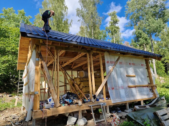 Person monterar svart tak på träkonstruktion, byggarbetsplats med byggmaterial, lummigt grönt träd i bakgrunden.