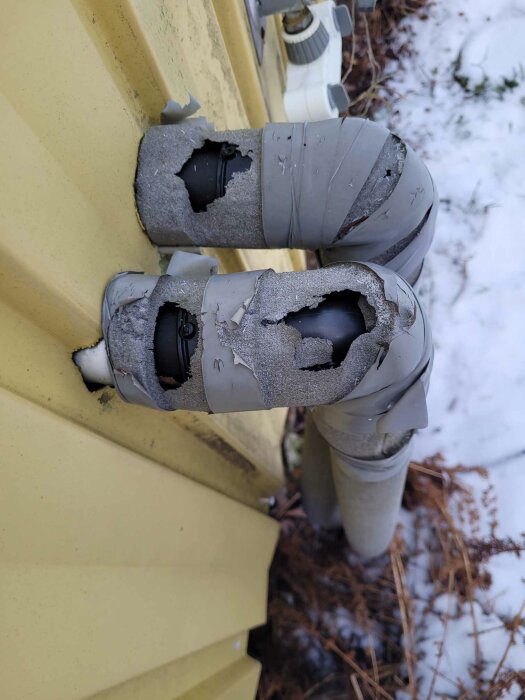 Isoleringsrör utomhus skadade, synliga sprickor, mot gul väggbeklädnad, snöaktig bakgrund.