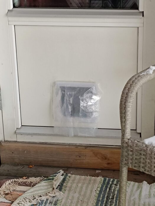 Ett fönster med en plastpåse tejpade över en öppning, trasig matta, en del av en korgstol framför.