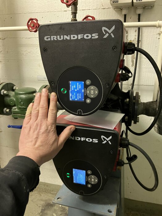 En hand vidrör en GRUNDFOS pump med digitala displaypaneler i ett maskinrum.