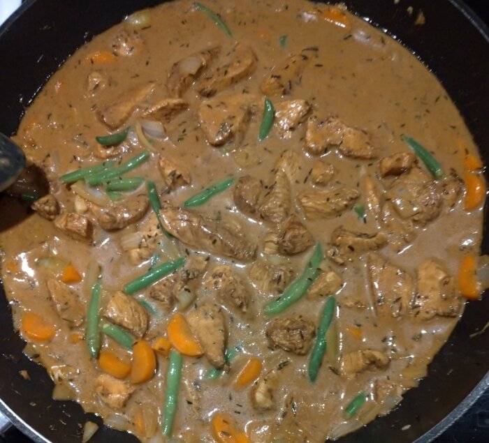 En stekpanna med gräddig kycklinggryta, morötter, gröna bönor och lök som tillagas.