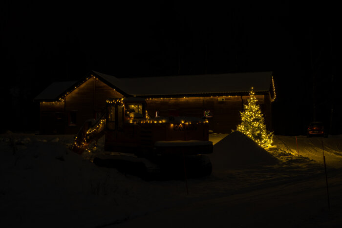 Hus och snöplog dekorerade med julbelysning under mörk kväll i snölandskap.