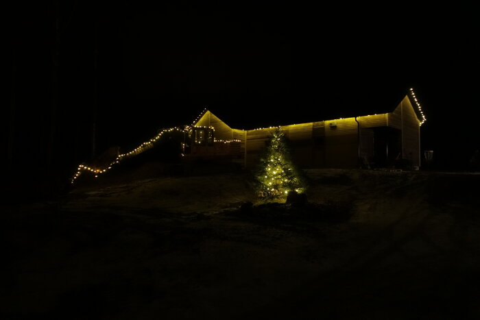 Ett hus med julbelysning i natten, upplyst julgran framtill, snöig mark.
