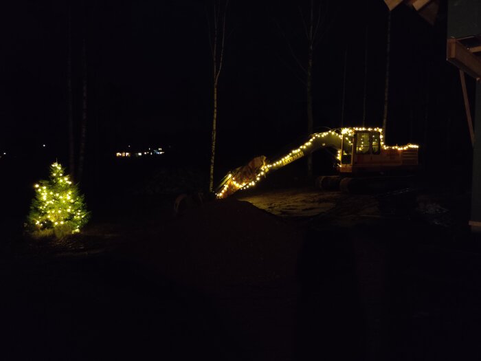 En grävmaskin och julgran dekorerade med ljusslingor i mörker. Ljus i bakgrunden. Natt. Utomhus. Festlig dekoration.