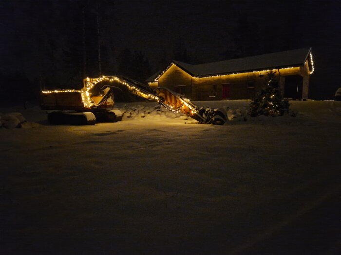 Ett hus och en grävmaskin dekorerade med ljusslingor i ett snötäckt landskap nattetid.