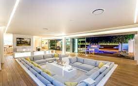 Modernt vardagsrum med stor soffa, infälld belysning, trägolv, glasväggar och utsikt över poolområde nattetid.