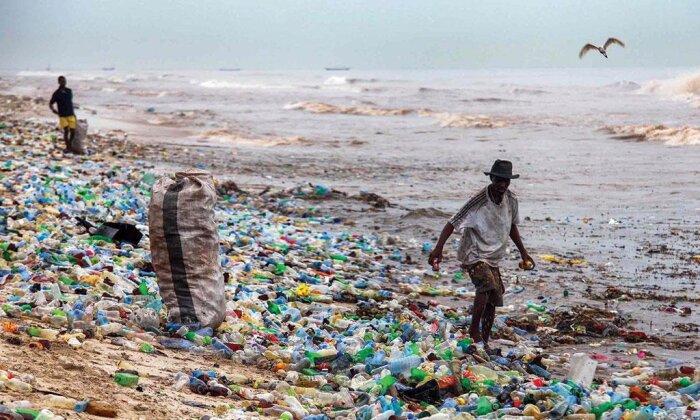 Strand överbelamrad med plastavfall, två personer verkar samla skräp, förorenat vatten, grå himmel, miljöproblem.