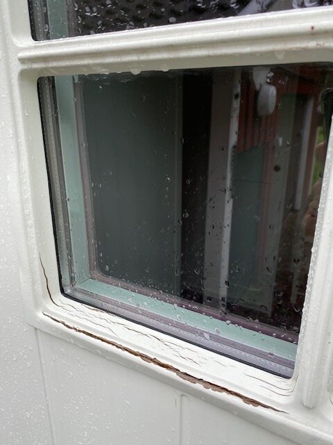 Närbild av ett fönster med regndroppar på en vit dörr, suddig bakgrund, möjlig fuktighetsskada längs karmen.