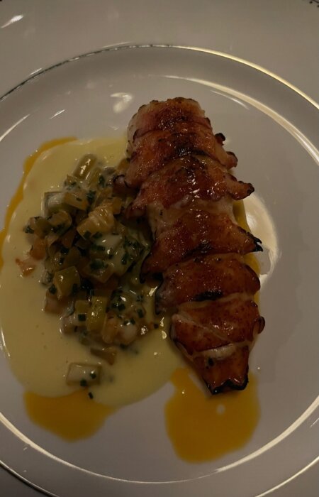 Grillat kött, möjligtvis kyckling, inlindad i bacon på vit tallrik med sås och grönsaker.